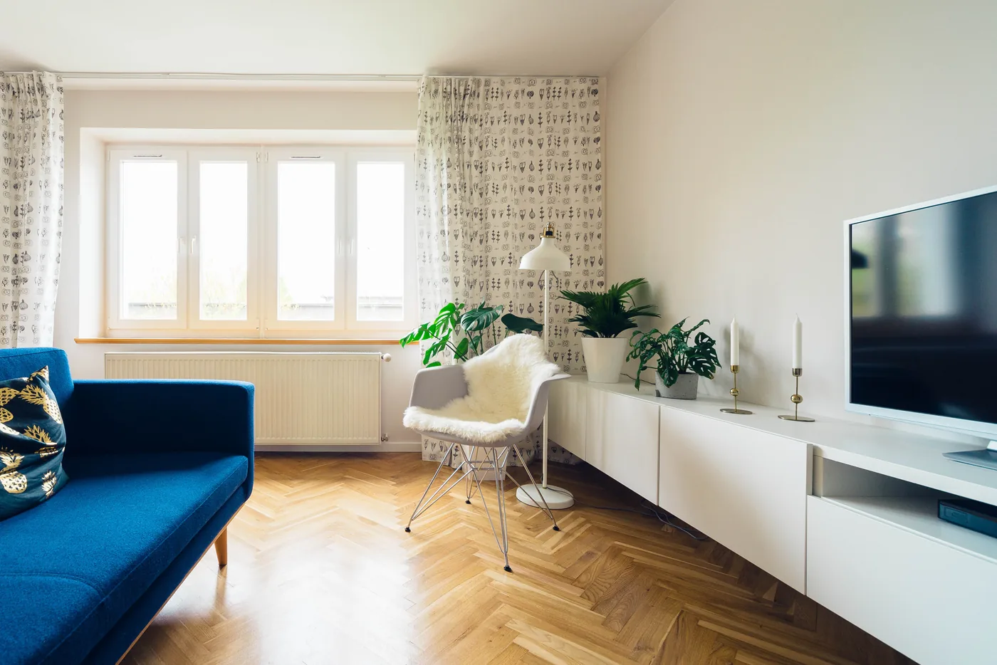 Ein blaues Sofa und ein weißer Sessel in einem hellen Wohnzimmer mit Pflanzen und einem ausgeschalteten Fernseher.