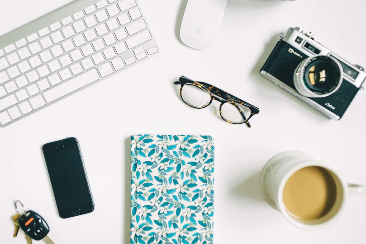 Originalfoto eines hellen Schreibtischs mit Kamera, Brille, Notizbuch, Smartphone und einer Tasse.
