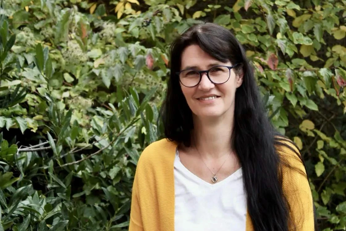 Tini Gerlach mit Brille und gelbem Cardigan vor grünen Blättern.