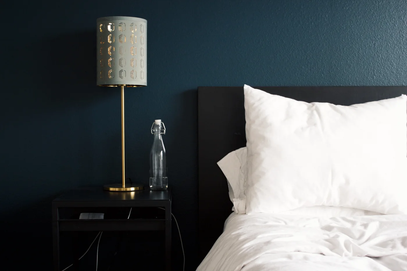 Ein dunkler Beistelltisch mit Lampe und Glasflasche neben einem Bett vor einer blauen Wand.